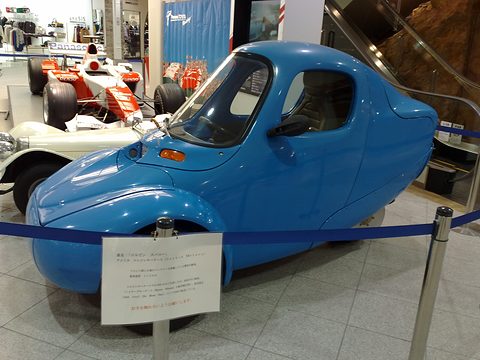 Blue teardrop-shaped three-wheeler behind a blue rope with an information placard in front of it, racing cars behind. Opens a larger view