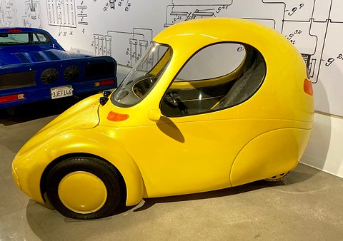 Side view of a yellow-gold teardrop single-seat car with a large door window and a disc-covered front wheel.