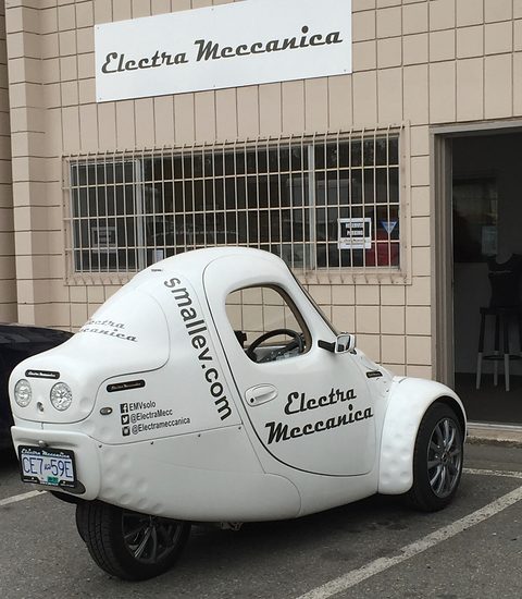 White single-seat three-wheeler lettered with a web address and social-media handles, parked outside a beige industrial unit. Opens a larger view