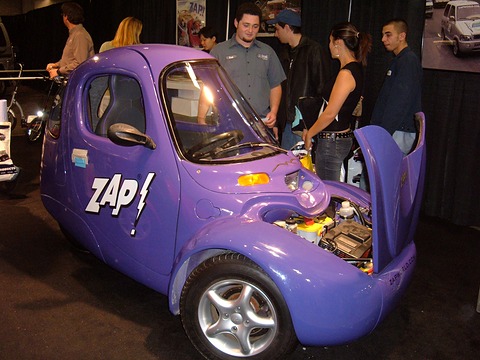 Purple three-wheeler with ‘ZAP!’ lettering and its nose panel lifted to show batteries and wiring, visitors behind.