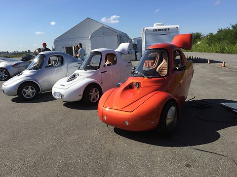 Two white and one red teardrop three-wheelers parked in a row on tarmac beside a white marquee under blue sky.