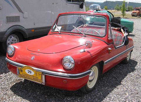 Small red doorless open roadster with round headlamps in the wings, chrome bumper strip and a yellow rally plate numbered 29, parked on gravel.