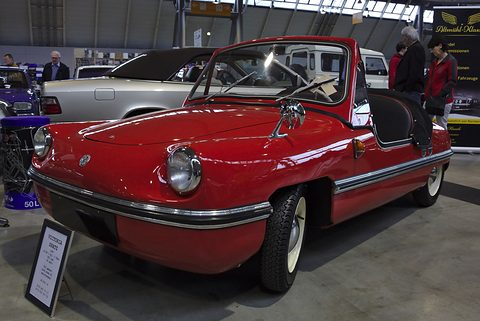 Glossy red doorless roadster with round headlamps, chrome bumper strip and a framed sign beside it, in an exhibition hall. Opens a larger view