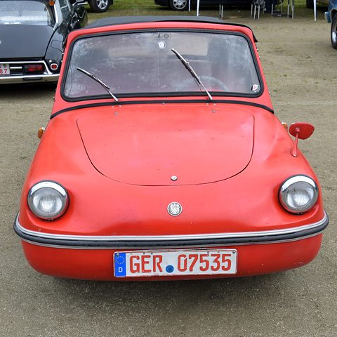Front of a red small roadster with a black hood, two round headlamps, a round badge and a German number plate, on a dusty field. Opens a larger view