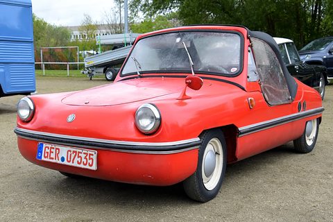 Weathered red doorless roadster with black cloth hood, clear sidescreens, round headlamps and chrome bumper strip, on a gravel field. Opens a larger view