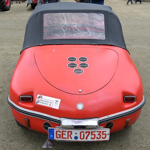 Rear of a red roadster with a black fabric hood and clear rear window, five oval vent grilles on the engine lid and a round badge.