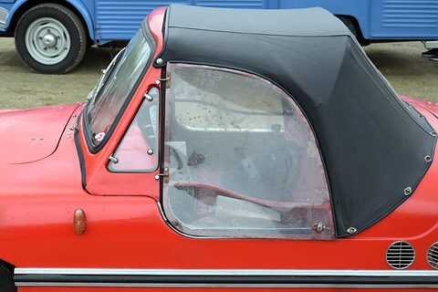 Side close-up of a red car showing a black fabric hood, a clear plastic side panel, a small hinged vent window and round chrome side vents.