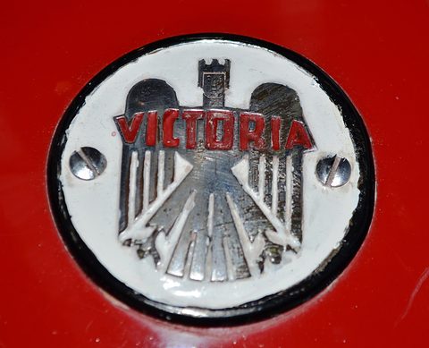 Close-up of a round white enamel badge with a chrome eagle and red VICTORIA lettering, set in red glass-fibre bodywork. Opens a larger view