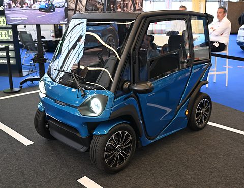 Blue two-seat microcar with a black frame, round LED headlamp and alloy wheels on a marked show floor. Opens a larger view
