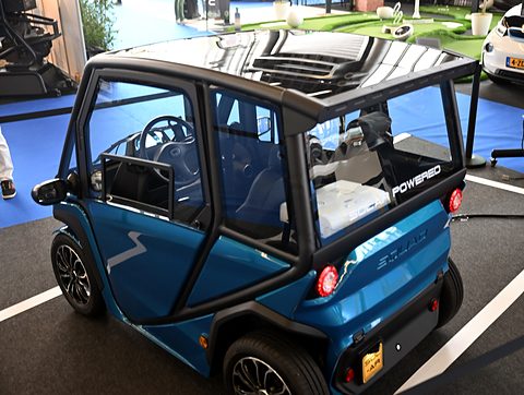 Blue and black open-framed two-seat microcar with a glossy solar roof on a dark show carpet. Opens a larger view