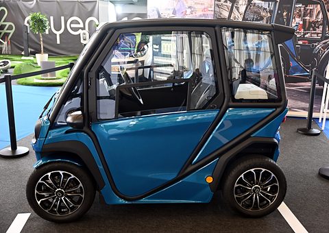 Side view of a blue two-seat microcar with a black frame, large windows and alloy wheels on a show carpet. Opens a larger view