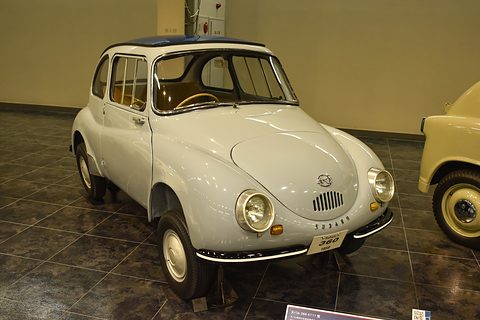 Pale grey small fastback saloon with a dark roof and round headlamps on a dark tiled floor, a cream car beside it. Opens a larger view