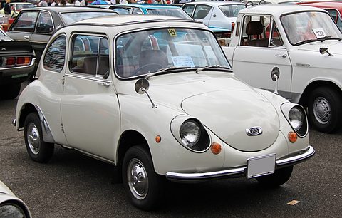 White small rounded saloon with front-hinged doors and a chrome bumper among other old cars in a car park. Opens a larger view