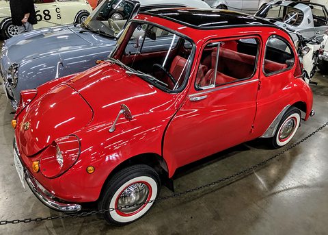 Red small rounded saloon with a black roof, white-wall tyres and red seats behind a chain, other cars around. Opens a larger view