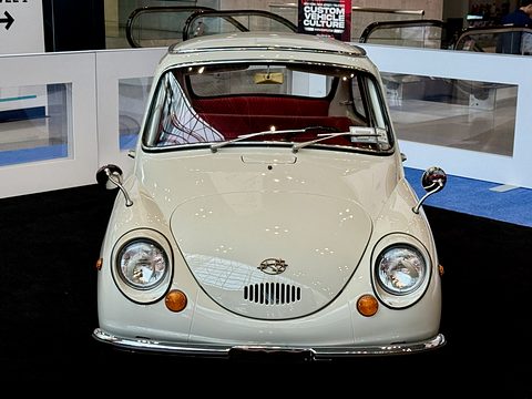 Front view of a cream small car with red seats, round headlamps and chrome mirrors on black carpet. Opens a larger view