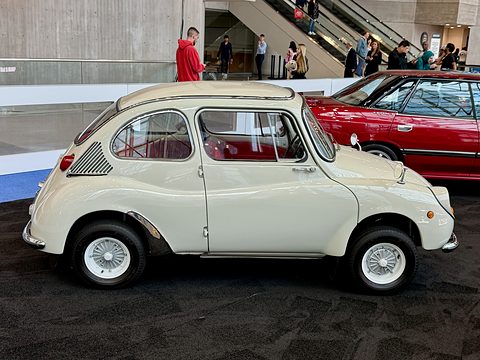 Side view of a cream small fastback car with chrome wheels and side air intakes, a red sports car behind. Opens a larger view