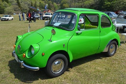 Bright green small rounded saloon with chrome bumpers and hubcaps on grass, a table and visitors behind. Opens a larger view
