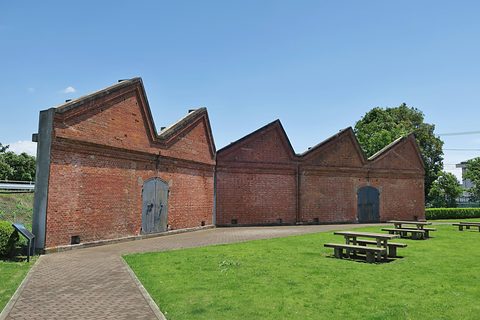 Long red-brick wall with saw-tooth gables and two blue doors beside a lawn with picnic tables under a blue sky.
