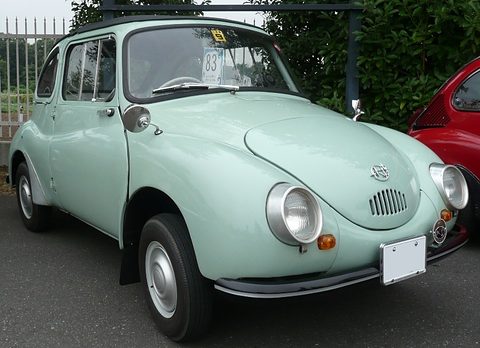 Pale green rounded small car with a dark fabric roof and chrome headlamp rims parked by a fence. Opens a larger view