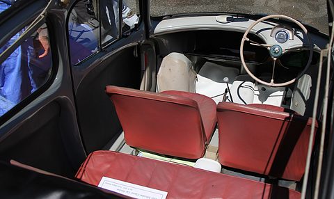 Looking down into a small car with red front seats, a red rear bench, a thin cream steering wheel and bare floor. Opens a larger view
