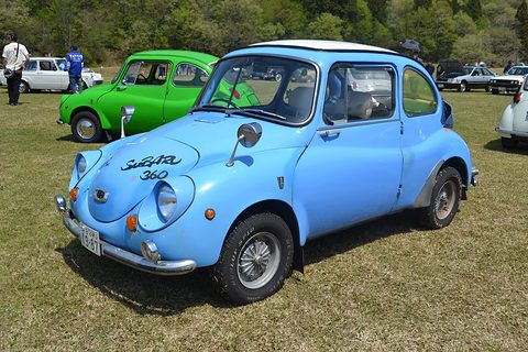 Light blue small rounded saloon with ‘Subaru 360’ painted on the bonnet, parked on grass beside a green car. Opens a larger view