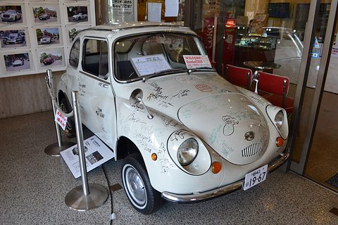 White small rounded car covered with handwritten signatures in black and green ink, photographs on the wall behind. Opens a larger view