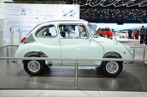 Side profile of a pale green small fastback car with a louvred rear panel and chrome wheels behind a rail. Opens a larger view