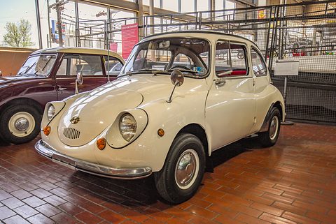 White small rounded saloon with front-hinged doors on a brick floor beside a maroon car, a mesh cage behind. Opens a larger view