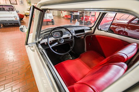 Open car door showing red vinyl seats, a black steering wheel and a small dashboard, a brick museum floor beyond. Opens a larger view