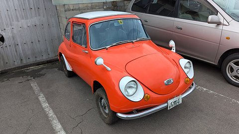 Orange small rounded saloon with a white roof and white headlamp surrounds in a car park beside a pink people carrier. Opens a larger view