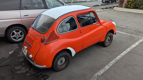 Rear three-quarter of an orange small car with a white roof and side air intakes, parked in a marked bay. Opens a larger view