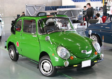 Green rounded small car with red roundels on its doors, chrome bumpers and spotlamps, parked in an exhibition hall. Opens a larger view