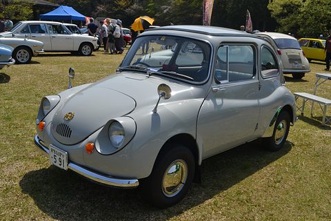Pale grey small rounded saloon with front-hinged doors and chrome hubcaps on grass, other old cars and gazebos behind. Opens a larger view