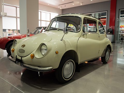 Cream small rounded saloon with white-wall tyres on a polished concrete floor, a red sports car behind. Opens a larger view