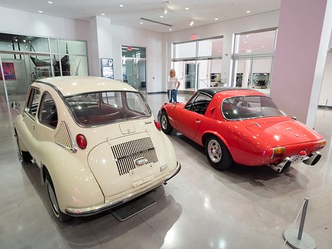 Rear of a cream small car with a slatted engine lid beside a red open sports car in a bright museum hall. Opens a larger view