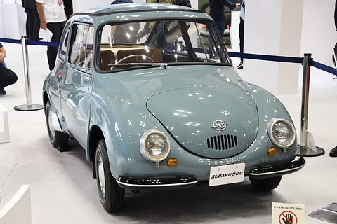 Grey-blue rounded small saloon with chrome-rimmed headlamps and a SUBARU 360 plate behind rope barriers. Opens a larger view