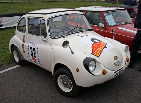 White small rounded saloon with a red rally plate on its bonnet and number 32 on its door, beside a red Mini. Opens a larger view
