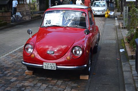 Front view of a red small car with round headlamps and a number card in its windscreen on a paved street. Opens a larger view