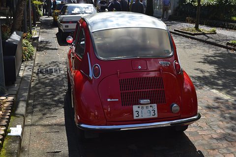 Red small rounded car with a silver roof and a slatted engine lid parked on a brick-paved street, other old cars behind. Opens a larger view