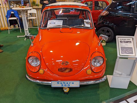 Front view of an orange small rounded car on green carpet beside an information stand and bistro tables. Opens a larger view