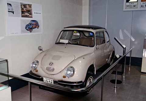 Beige small rounded saloon with a dark roof behind a curved rail, posters of the car on the wall behind. Opens a larger view