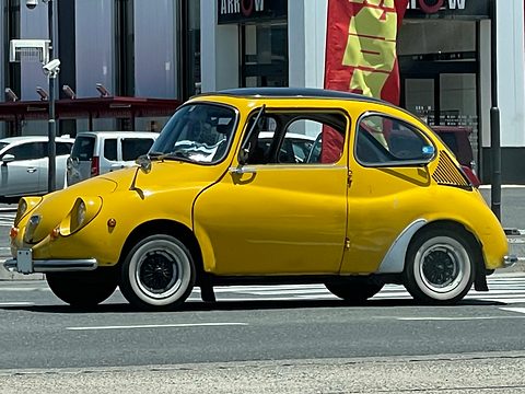 Side view of a yellow small rounded car with white-wall tyres crossing a road in front of shops.
