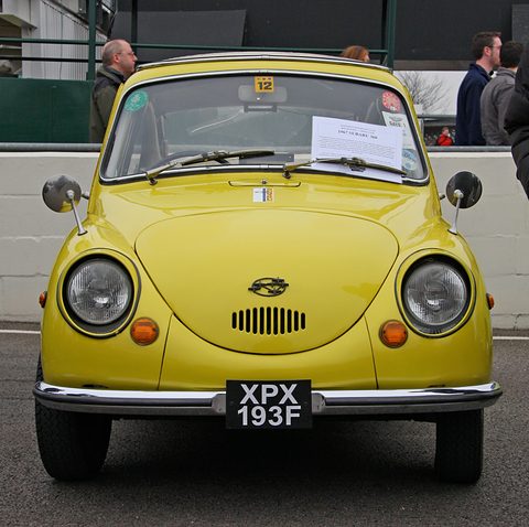 Front view of a yellow small rounded car with a black UK number plate and a document in its windscreen. Opens a larger view