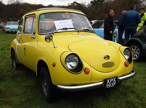 Yellow small rounded saloon with a UK plate and rally badges parked on wet grass, people chatting behind. Opens a larger view