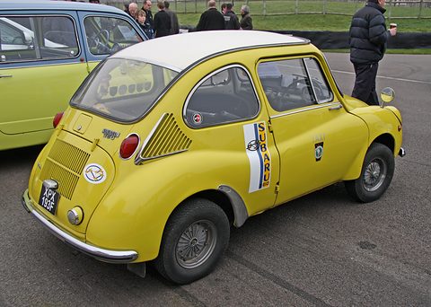Rear three-quarter of a yellow small car with a white roof, slatted engine lid and blue-and-white Subaru door stripe. Opens a larger view
