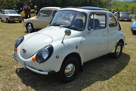 White small rounded saloon with black-ringed headlamps and chrome tube bumpers on grass, visitors behind. Opens a larger view