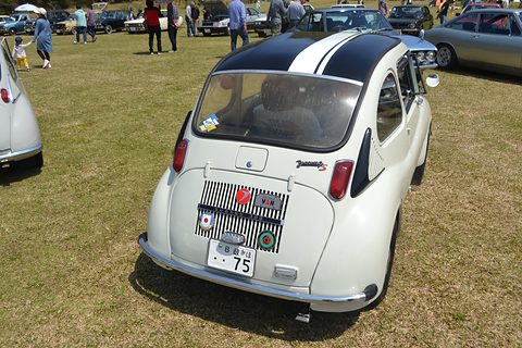 White small car seen from behind and above with two black stripes over its roof and badges on the engine lid. Opens a larger view