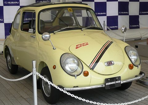 Pale yellow small rounded car with a black and red stripe and red SS letters on its bonnet behind a chain barrier. Opens a larger view