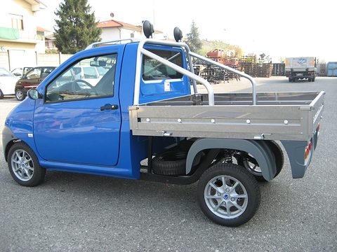 Small blue modern pick-up with alloy wheels, a roll bar and an aluminium drop-side load bed, in a yard. Opens a larger view