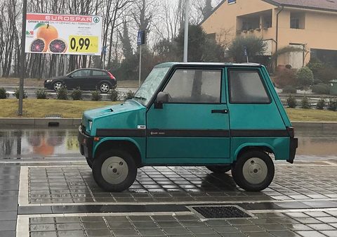 Green boxy microcar with four wheels and a black side stripe, parked on wet paving with a supermarket billboard behind.